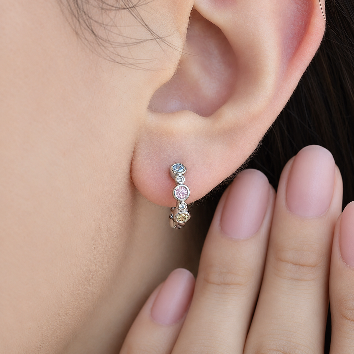 Silver-tone hoop with alternating small and larger pastel-coloured round stones worn on an earlobe.