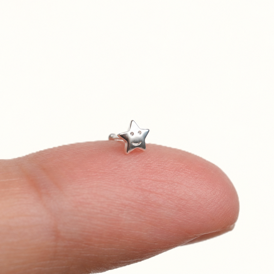 Silver-coloured smiling star nose stud resting upright on a fingertip.