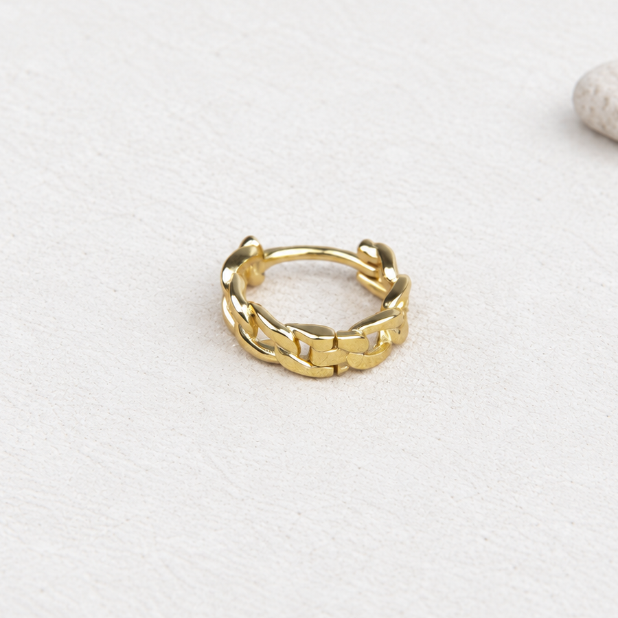 Gold-coloured hoop earring with a raised chain-link pattern resting on a pale textured surface beside a pebble.