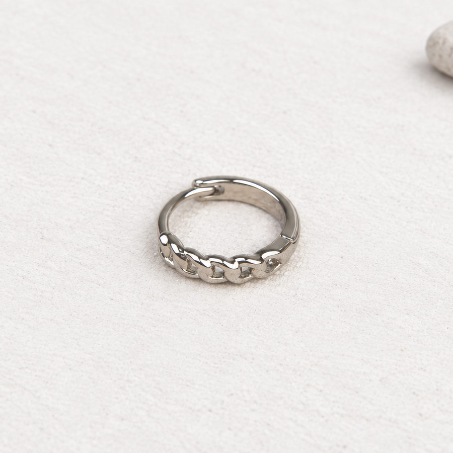 Silver-coloured hoop earring with a chain-link pattern resting on a pale textured surface beside a pebble.
