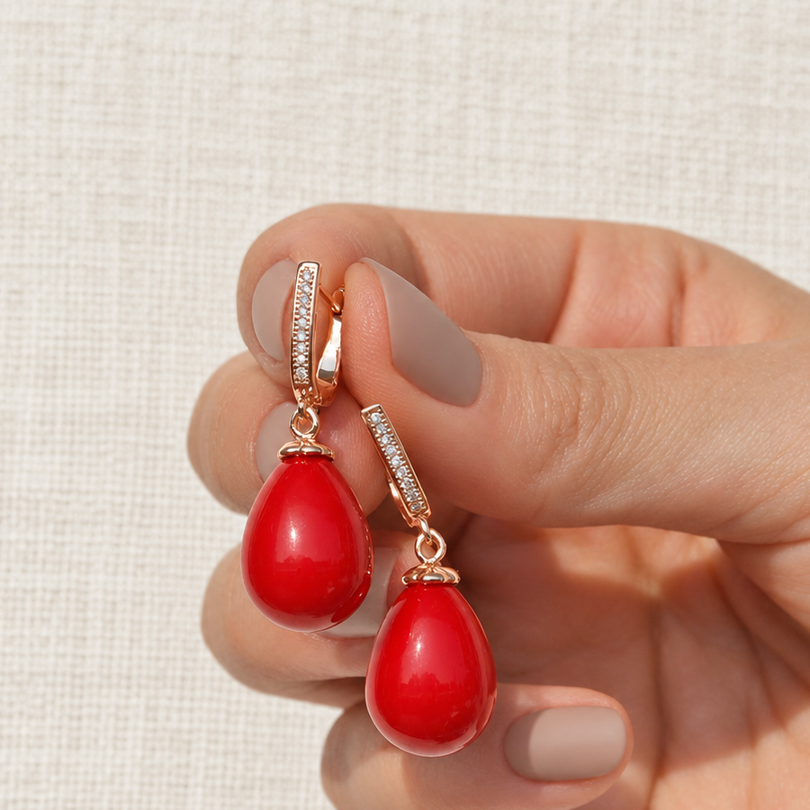 Two clear-stone earrings with glossy red teardrops held between fingers.