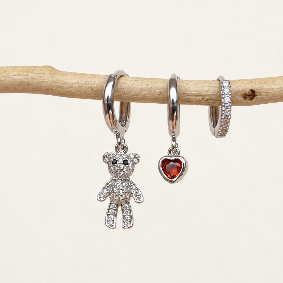Three silver-tone hoops hanging from a branch, with a clear-stone teddy bear, red heart and stone-lined hoop.
