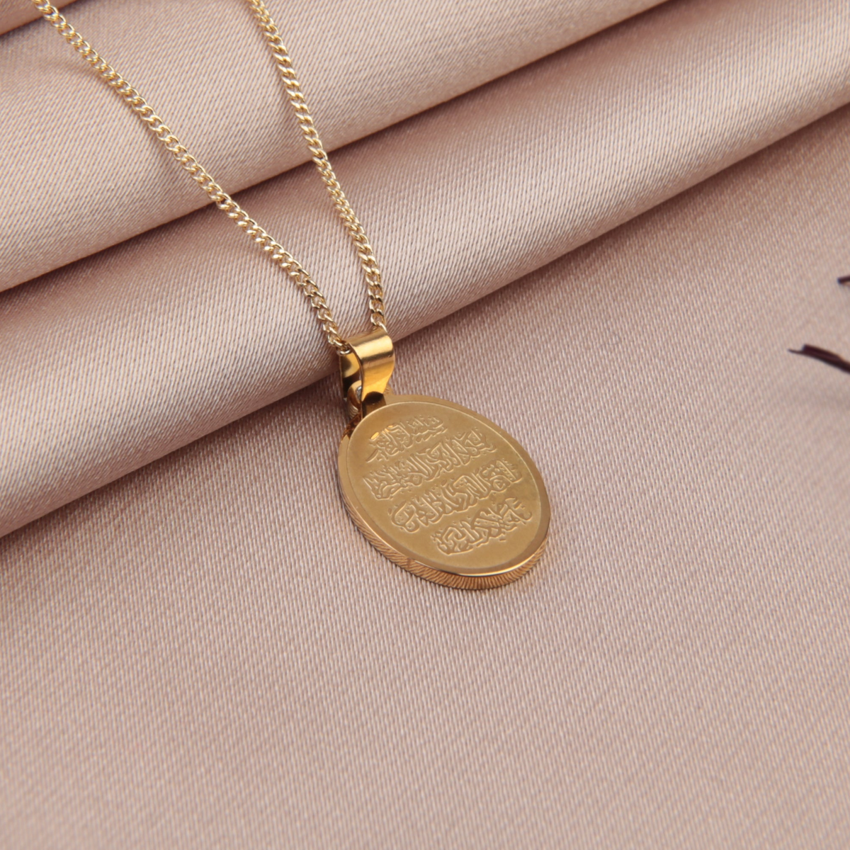 Gold-coloured oval Arabic calligraphy pendant and fine chain on beige fabric beside dried red petals.