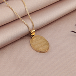 Gold-coloured oval Arabic calligraphy pendant and fine chain on beige fabric beside dried red petals.