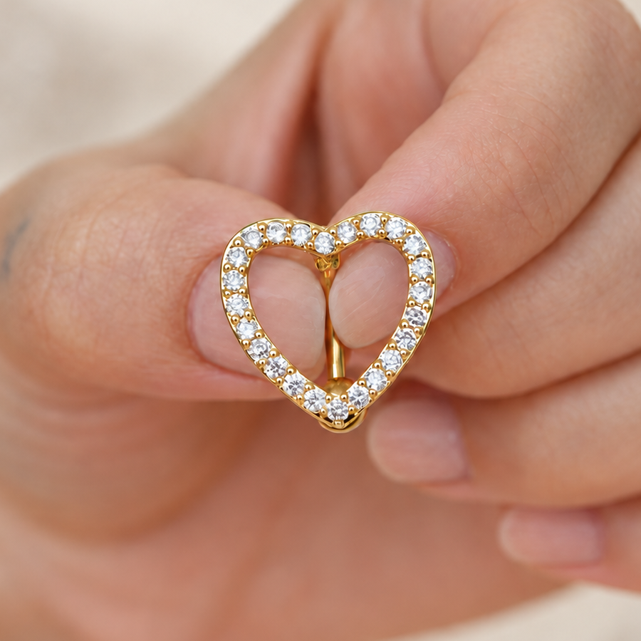 Gold-tone open-heart belly bar outlined with clear stones, held between fingers.