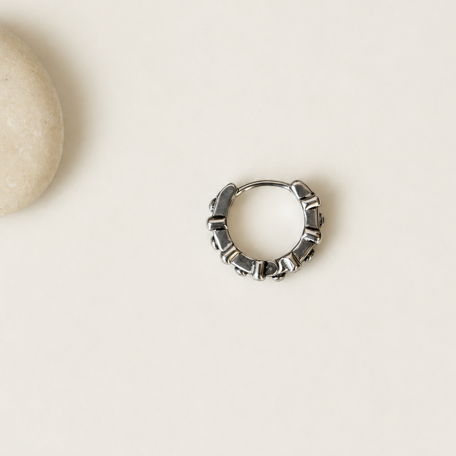 Closed dark silver-tone hoop earring with raised geometric details on a cream surface.