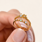 Gold-tone double-band ring with a small flower and pastel-coloured stones held between fingers.