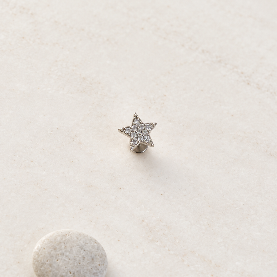 Silver-tone clear-stone star piercing stud on a pale surface beside a small pebble.