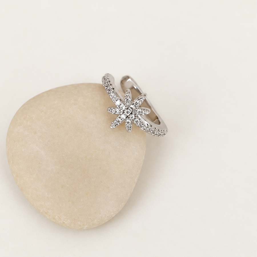 Silver-tone ear cuff with a clear-stone starburst and slim stone-lined band resting on a pale pebble.