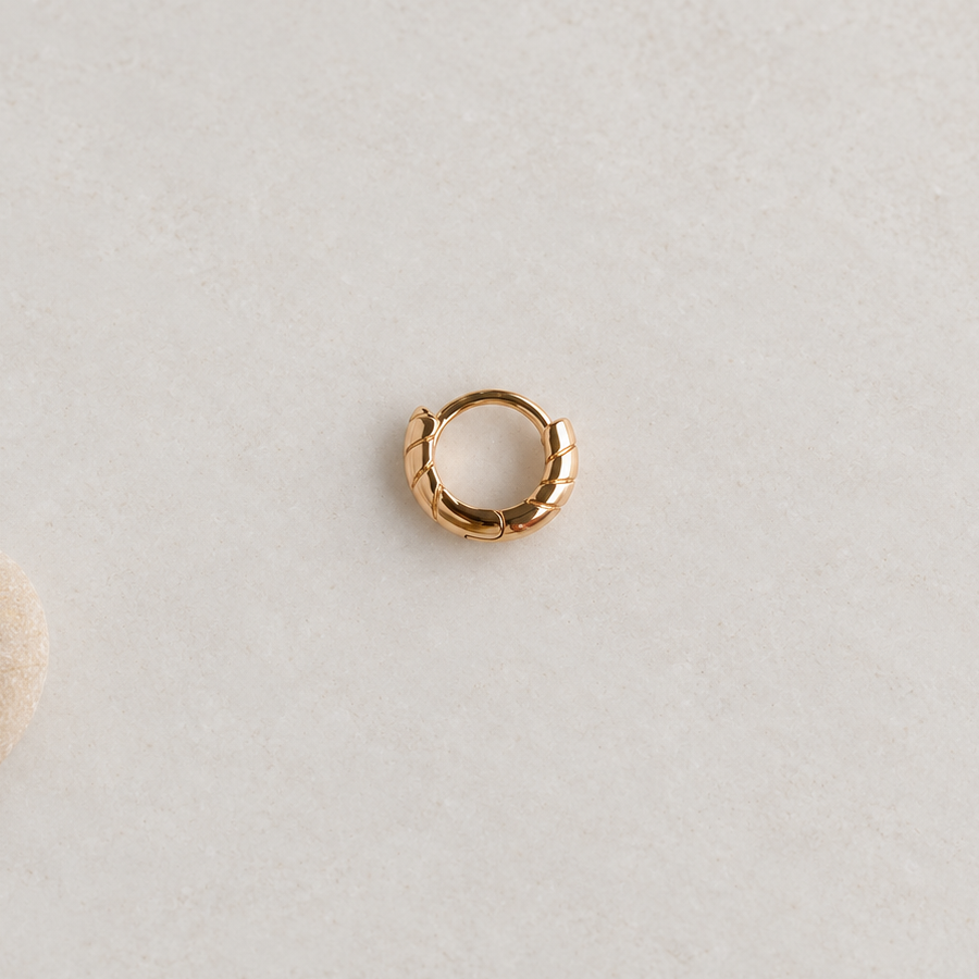 Gold-tone grooved hoop with its clasp closed on a pale surface beside a pebble.