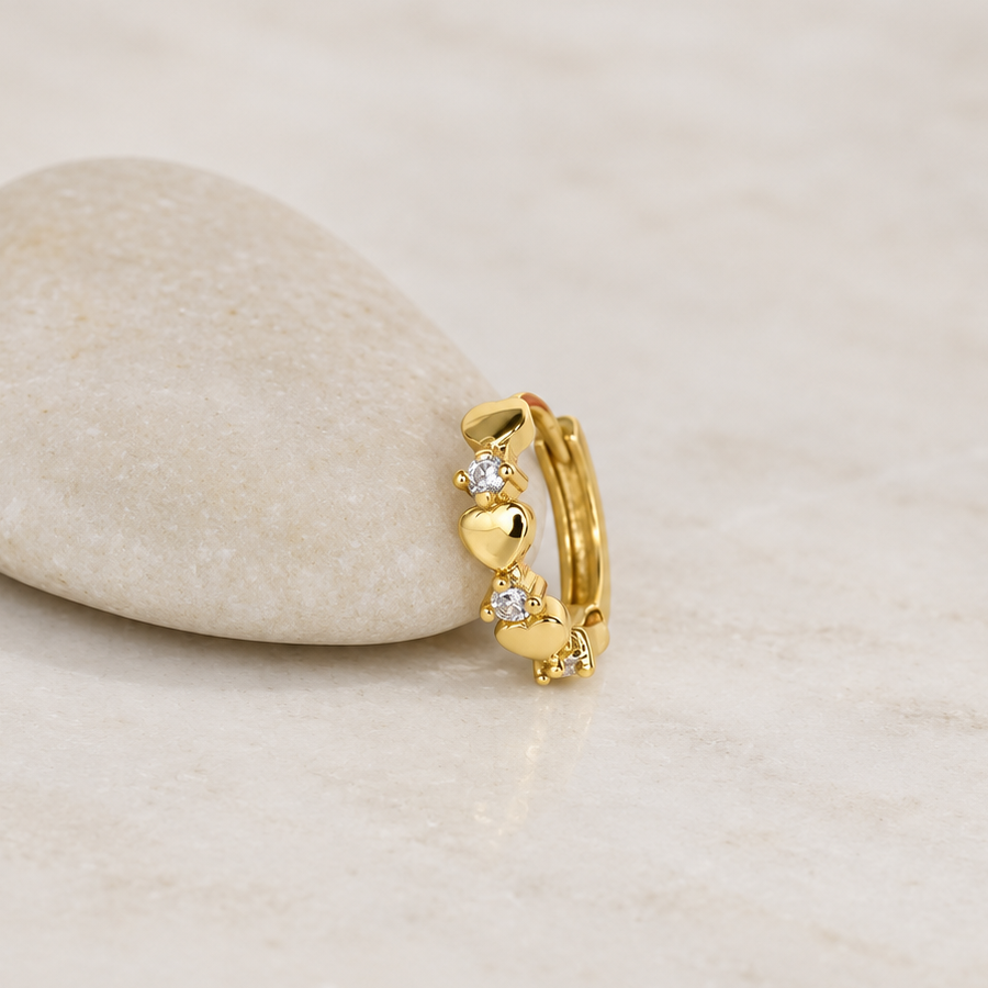 Gold-coloured hoop with alternating polished hearts and clear stones leaning against a pale pebble.