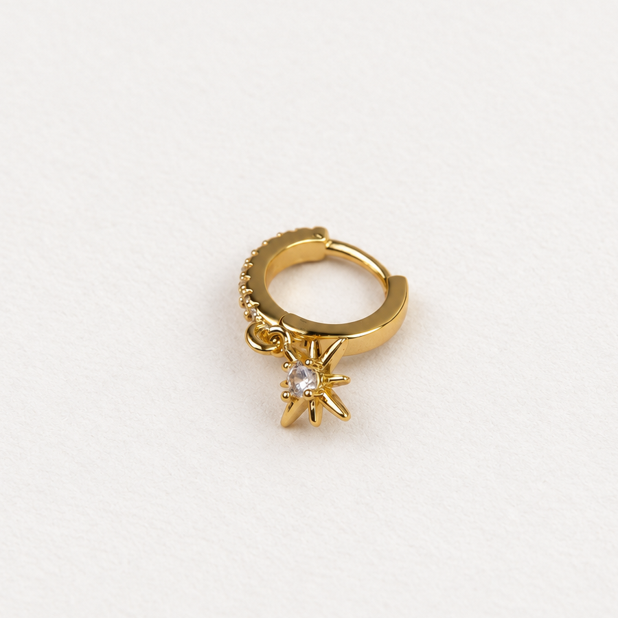 Gold-tone hoop with clear stones and a dangling starburst charm lying on a white surface.