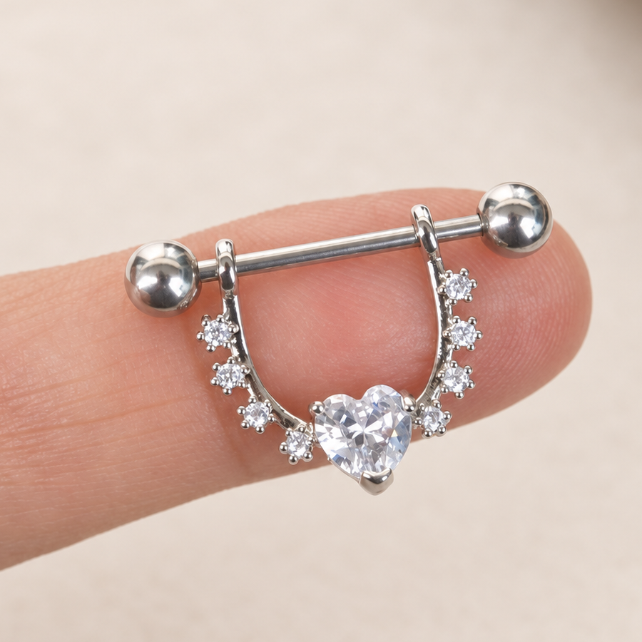 Silver-coloured nipple barbell with a clear heart stone and curved stone-set shield balanced on a fingertip.