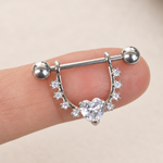 Silver-coloured nipple barbell with a clear heart stone and curved stone-set shield balanced on a fingertip.