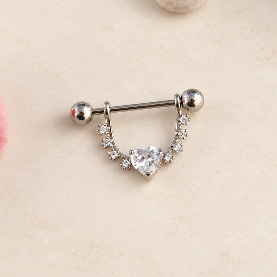 Silver-coloured nipple barbell with a clear heart stone and curved stone-set shield on a pale surface.