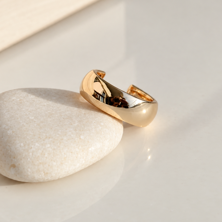 Wide gold-coloured curved ear cuff leaning against a white pebble on a reflective pale surface.