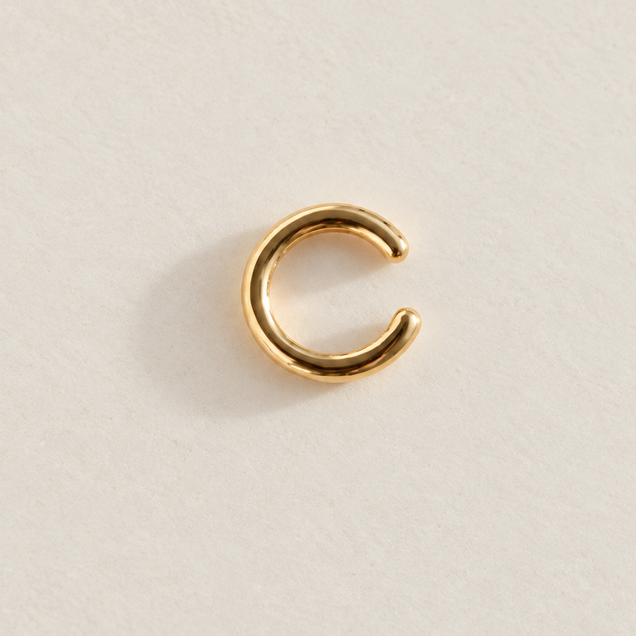 Gold-tone curved ear cuff with an open gap, laid flat on a cream surface.