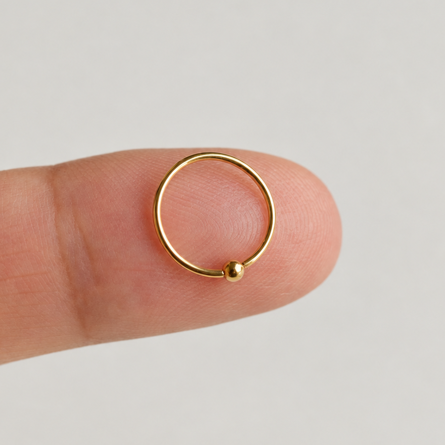 Gold-tone nose ring with a small ball resting on a fingertip.