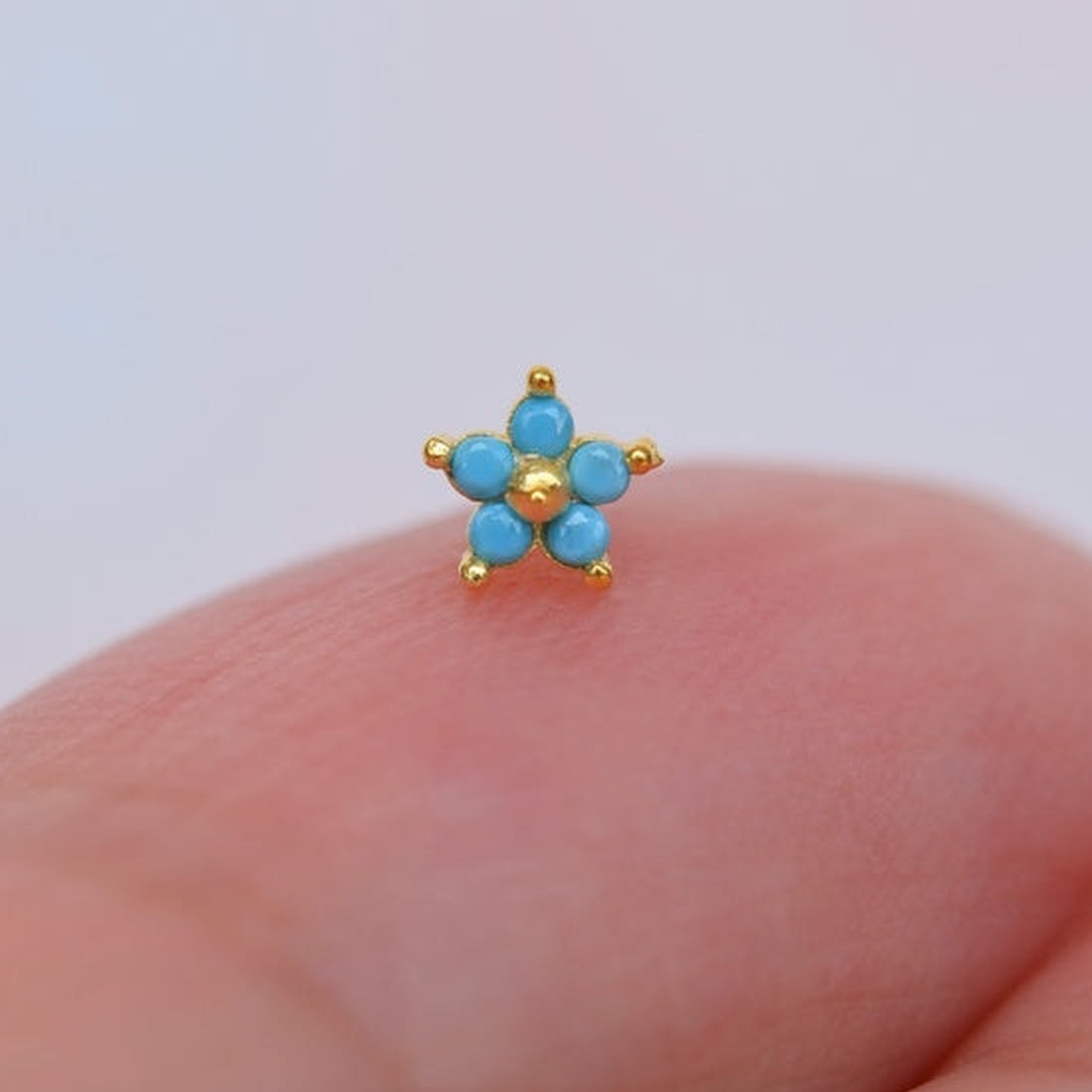 Gold-tone nose stud with five turquoise-blue flower petals and a round centre, displayed on a fingertip.