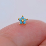 Gold-tone nose stud with five turquoise-blue flower petals and a round centre, displayed on a fingertip.