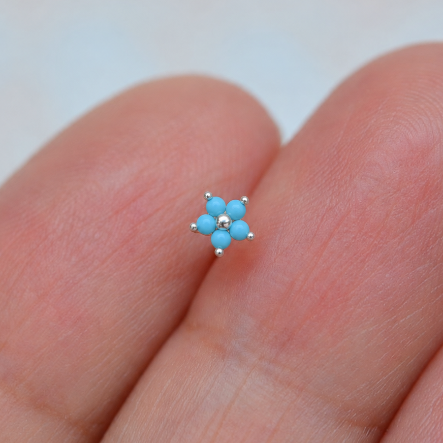 Silver-tone nose stud with five turquoise-blue flower petals and a round centre, displayed on fingers.