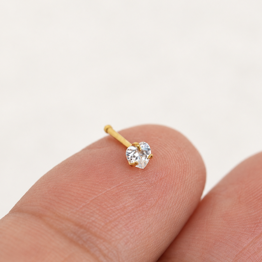 Gold-tone nose stud with a clear heart-shaped stone and straight post resting on a fingertip.