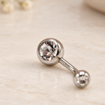 Silver-tone curved belly bar with two clear-stone ball ends lying on a pale surface.