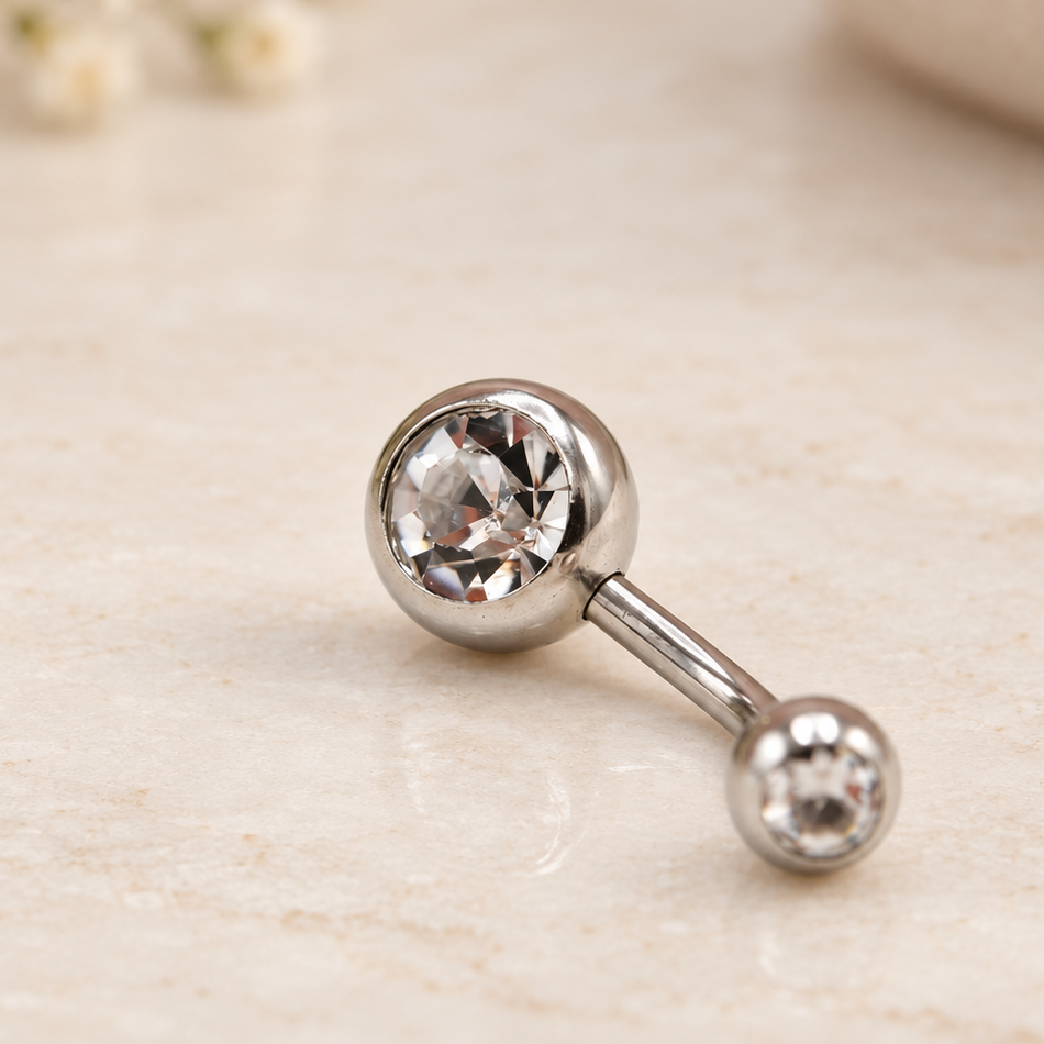Silver-tone curved belly bar with two clear-stone ball ends lying on a pale surface.