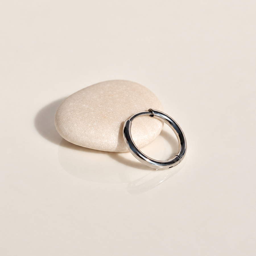 Polished silver-tone hoop earring leaning against a pale pebble on a cream surface.