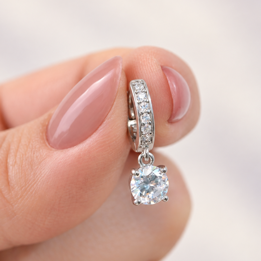 Silver-tone stone-set hoop earring with a dangling round clear stone, held between fingertips.