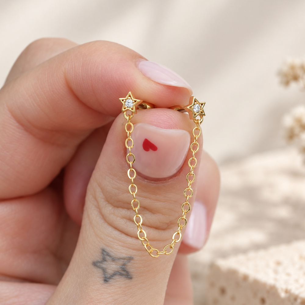 Two gold-tone star studs with clear centres linked by a dangling chain, held between fingertips.