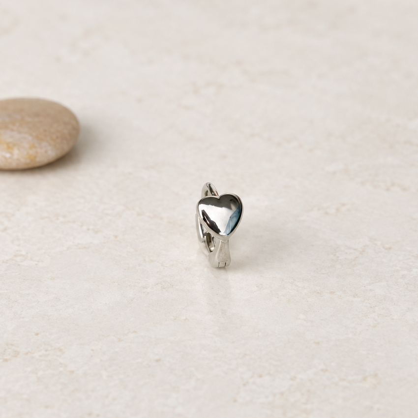 Silver-tone heart-front hoop earring standing on a pale textured surface beside a pebble.